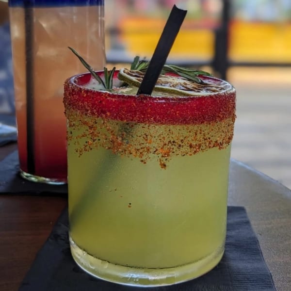 A vibrant green cocktail with a red and brown spiced rim, garnished with lime and rosemary.