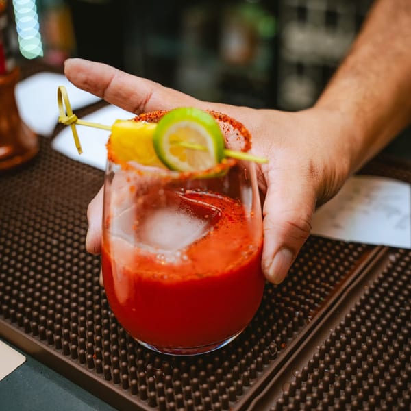 A hand holding a red Mexican cocktail garnished with lime, lemon, and chili powder.