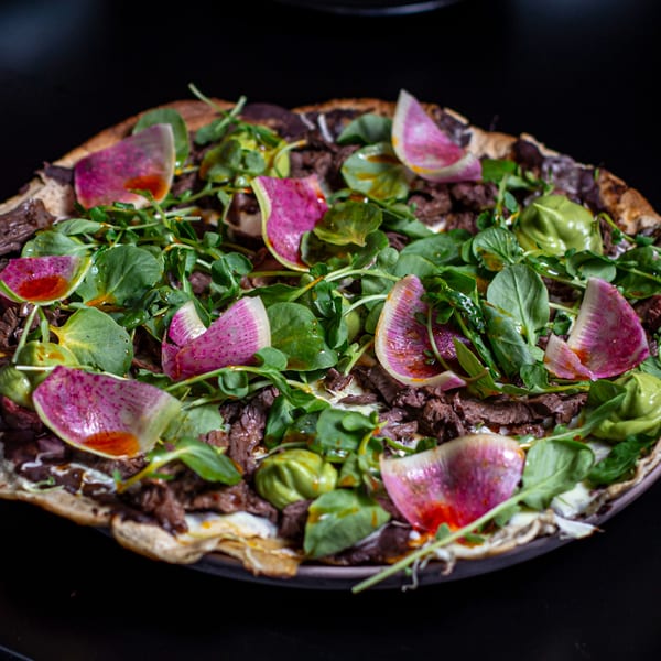 Chicatana's Mexican pizza with sliced beef, green leaves, avocado, and watermelon radish on a dark background.