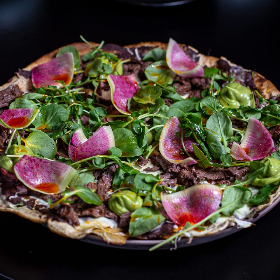 Chicatana's Mexican pizza with sliced beef, green leaves, avocado, and watermelon radish on a dark background.