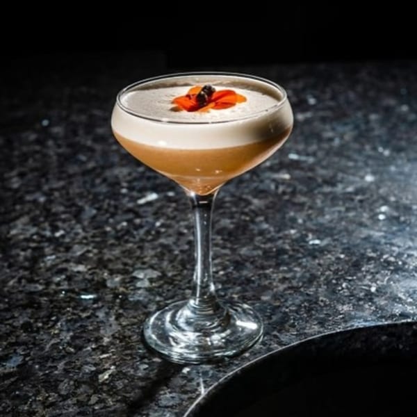 Chicatana cocktail with foamy top and orange garnish on a dark, speckled counter.
