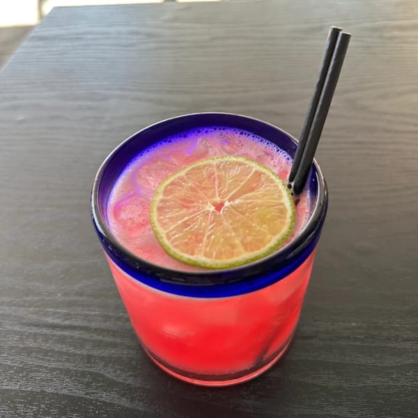 Vibrant pink cocktail with a lime slice and two black straws in a blue-rimmed glass.