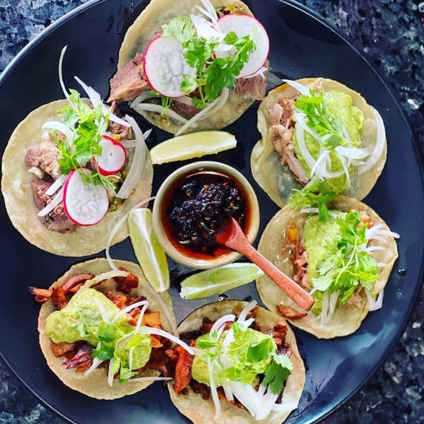 A black plate holds six Chicatana tacos with various fillings, guacamole, cilantro, onions, and radishes.